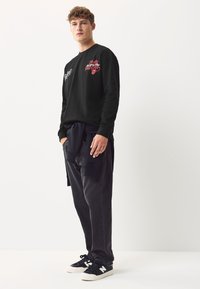 Sort langærmet skjorte med hvide og røde broderier, kombineret med mørkegrå bukser og sorte sneakers med hvide logo-accenter.