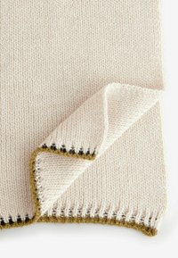 Gestrickte, cremefarbene Decke mit einer umgeschlagenen Ecke, die eine strukturierte Kante zeigt, akzentuiert mit braunen und senfgelben Details.