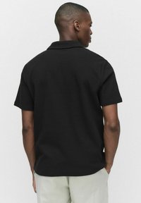 Svart kortärmad skjorta i texturerad tyg med krage, som visar en avslappnad passform och sömlösa axlar. Baksidan framhäver kragdesignen.