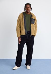 Tan fleecejakke med en sort lynlåslomme, grøn sweater under, parret med marineblå fløjlssystem og hvide sneakers.