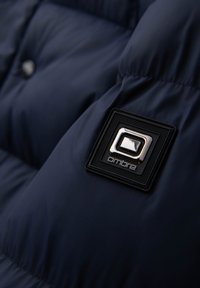 Granatowa kurtka puchowa z błyszczącą czarną metką z srebrnymi akcentami. Teksturowana powierzchnia pokazuje pikowany wzór.