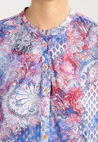 Colorful patterned blouse featuring a mixed paisley design in shades of blue, pink, and red. It has a stand collar and orange buttons.