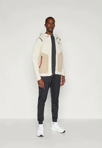 Gaiši krēmkrāsas jaka ar rāvējslēdzēju, ar bēšām paneļām un sarkaniem akcentiem, kopā ar tumši zilām biksēm un baltām sporta kurpēm. Gluda tekstūra un pieguļošs dizains.