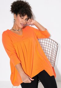 Oranges, V-Ausschnitt-Oberteil mit Dreiviertelärmeln und gefälteltem Design. Kombiniert mit schwarzen Leggings, vor einem weißen Hintergrund.