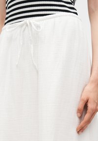 Γυναίκα φοράει λευκό παντελόνι με κορδόνι, συνδυασμένο με μαύρο-λευκό ριγέ τοπ, το χέρι ακουμπάει στο πλάι της.