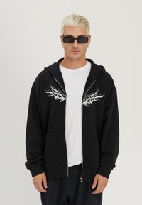 Młody mężczyzna z krótkimi blond włosami, noszący czarne okulary przeciwsłoneczne, czarną bluzę z kapturem na zamek z białymi wzorami tribal oraz białą koszulkę na tle jednolitego tła.