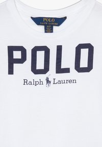 Camiseta blanca con gran texto negro "POLO", texto más pequeño "Ralph Lauren" y un logo de jugador de polo debajo de la etiqueta del cuello.