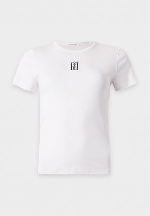 Giovane donna in piedi, indossa una t-shirt bianca a maniche corte con un piccolo logo e jeans larghi azzurri chiari con sandali neri.