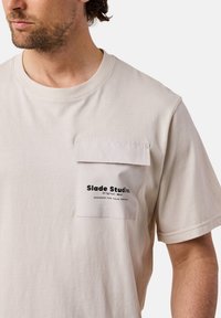 Beige bomullströja med en bröstficka med en vit klaff och svart tryckt logotyp. Kortsleeve och rund halsringning.