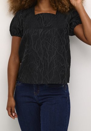 Kobieta ubrana w czarną, teksturowaną bluzkę z krótkim rękawem i kwadratowym dekoltem oraz ciemnoniebieskie dżinsy, stojąca na jednolitym tle.