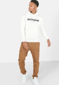 Witte hoodie met zwarte opdruk "SIXTH JUNE PARIS"; bruine cargo broek met meerdere zakken; witte sneakers. Schone, casual outfit.