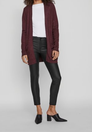 Femme portant un cardigan bordeaux ouvert, un haut blanc, un pantalon noir effet cuir et des mules noires à talons, debout devant un fond uni.
