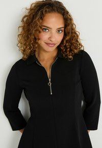 Robe noire avec une fermeture éclair sur le devant, dotée d'un col et de manches trois-quarts. Tissu lisse, design évasé, avec un tirette en forme de cœur.
