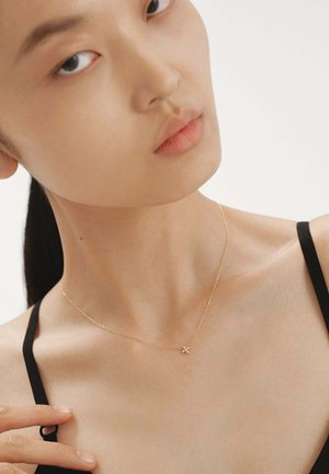 Collar de oro con un pequeño colgante en forma de flor sobre una cadena fina, llevado en un cuello desnudo con piel clara y suave contra un fondo neutro.