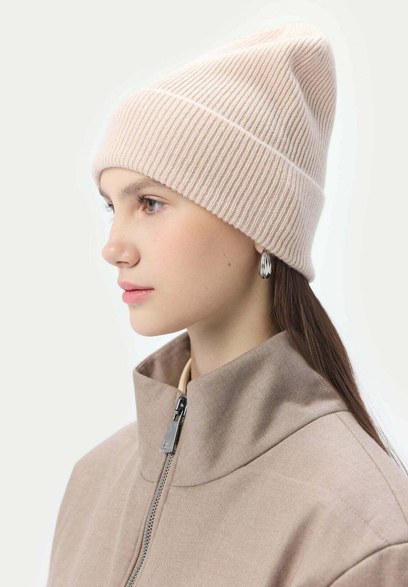 Stickad beige mössa med ribbat mönster, med en vikt kant och subtila sömdetaljer, bärs med en högkragad beige jacka.
