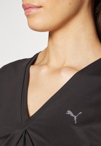 Sort svart v-hals topp med en innsnørt design, med en subtil grå Puma-logo på venstre brystområde. Glatt stoff, tettsittende fasong.