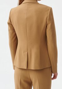 Op maat gemaakte blazer in camel kleur, gemaakt van gladde stof. Heeft een getailleerd silhouet, een gelapelde kraag en een split in het midden aan de achterkant.