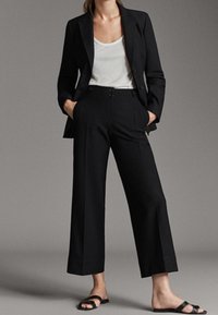 Zwarte op maat gemaakte blazer met een enkele knoop, gecombineerd met hoge zwarte wijde broek. Gladde stof, minimalistisch ontwerp, gedragen met platte sandalen.