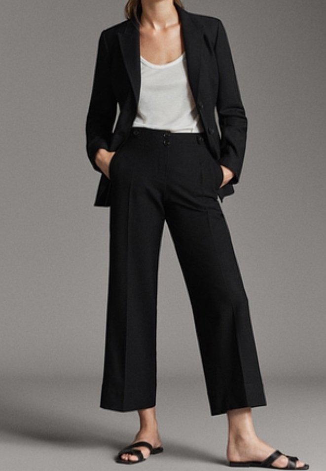 Zwarte op maat gemaakte blazer met een enkele knoop, gecombineerd met hoge zwarte wijde broek. Gladde stof, minimalistisch ontwerp, gedragen met platte sandalen.