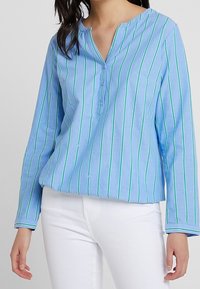 Chemise à manches longues bleu clair avec des rayures verticales vertes, col boutonné et coupe décontractée, assortie à un pantalon blanc.