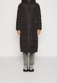 Black puffer coat with a hood, zip closure, and side pockets; features quilted design and a longer length, paired with gray jeans and silver shoes.