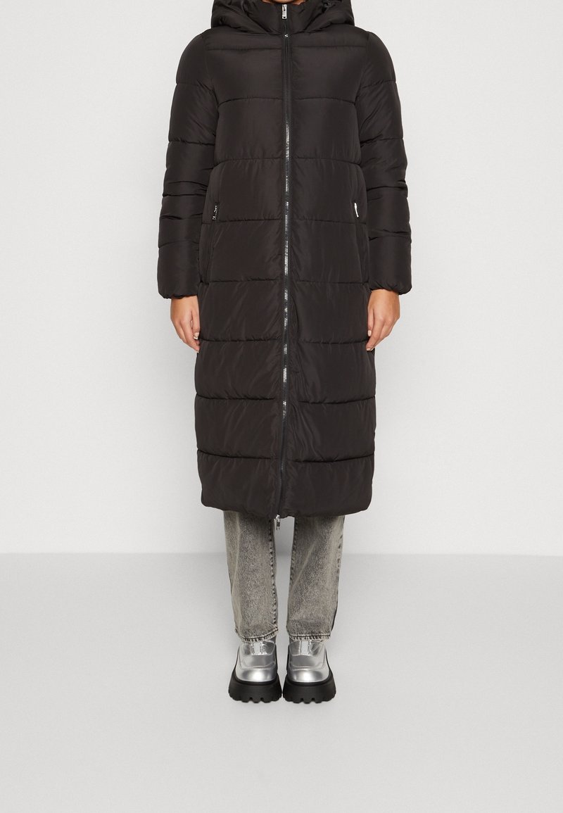 Black puffer coat with a hood, zip closure, and side pockets; features quilted design and a longer length, paired with gray jeans and silver shoes.