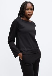 Črna majica z dolgimi rokavi iz gladkega materiala z okroglim izrezom. Na pasu je teksturiran trak z imenom blagovne znamke "HUGO" v črni barvi.