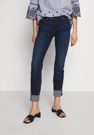 Dark blue skinny jeans with a cuffed hem and contrasting stitched detail, paired with black slip-on sandals. Cotton-blend material.