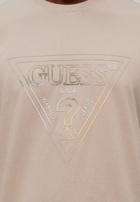 Μπεζ πουκάμισο με μεταλλικό χρυσό και λευκό λογότυπο Guess, που περιλαμβάνει ένα τρίγωνο και ένα ερωτηματικό, στο κέντρο του στήθους.