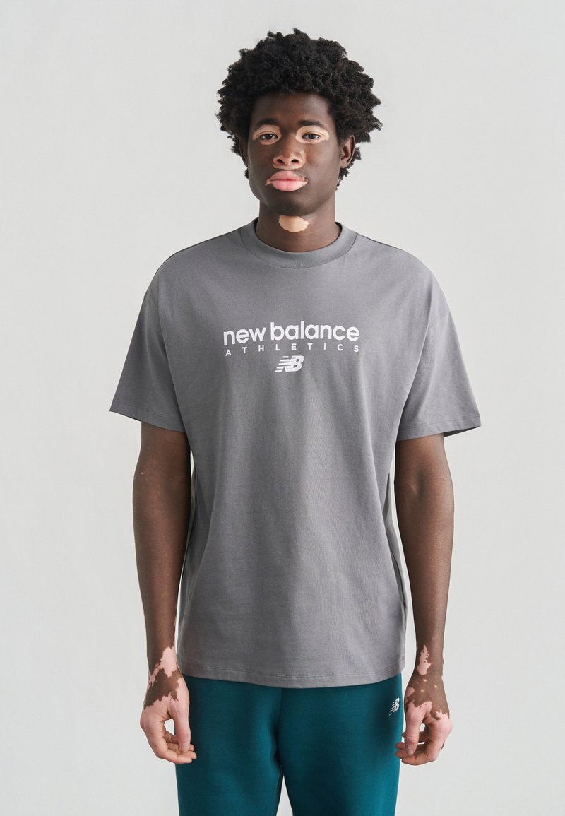 Γκρι βαμβακερό μπλουζάκι με κοντά μανίκια, με λευκό λογότυπο «new balance» και το κείμενο «ATHLETICS» μπροστά. Ομαλή υφή, άνετη εφαρμογή.