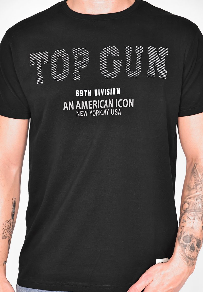 Svart t-shirt med stor vit och prickig text som säger "TOP GUN", "69TH DIVISION" och "AN AMERICAN ICON" med en mjuk struktur.
