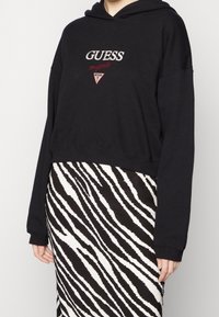 Svart kort hoodie med broderad "GUESS"-logotyp och röda detaljer, kombinerad med en svart och vit zebra-mönstrad kjol. Slät tygkvalitet.