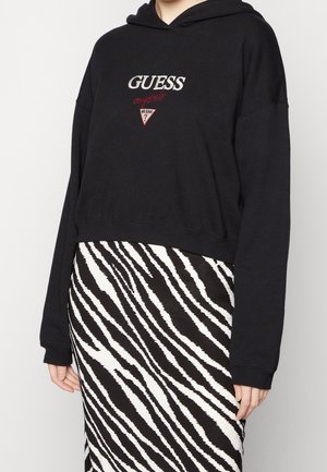 Haut à capuche noir court avec logo "GUESS" brodé et accents rouges, associé à une jupe à motif zèbre noir et blanc. Texture de tissu lisse.