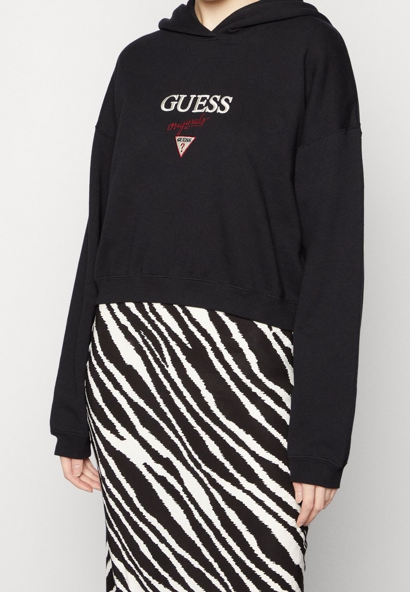 Svart kort hoodie med broderad "GUESS"-logotyp och röda detaljer, kombinerad med en svart och vit zebra-mönstrad kjol. Slät tygkvalitet.