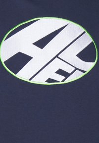 T-shirt blu con un grande logo ricamato bianco contornato di verde, che mostra un design grafico audace.