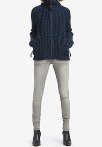 Tmavě modrý pletený svetr na zip s texturálním vzorem, vysokým límcem, bočními kapsami a nastavitelnými šňůrkami. Kombinován s světle šedými úzkými džíny.