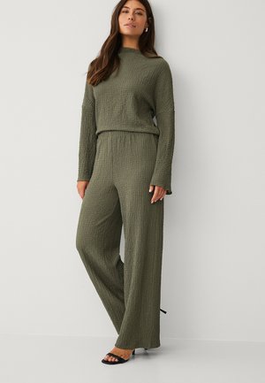 Kvinde iført olivengrøn tekstureret top med lange ærmer og bukser med vide ben, stående foran en ensfarvet lysegrå væg, iført sorte sandaler med hæl.