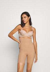 Naadloze shapewear shorts in huidskleur met een gladde textuur en hoge taille, voorzien van een rekbaar pasvorm en lengte tot halverwege de dij. Boven een bh met kantaccent.