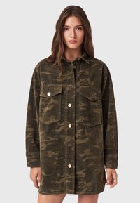Junge Frau, die eine übergroße olivgrüne Kordjacke im Camouflage-Muster mit vorderen Knöpfen und zwei aufgesetzten Brusttaschen trägt, steht vor einem schlichten Hintergrund.