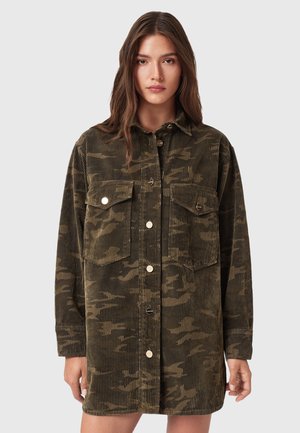 Young woman wearing an oversized olive green corduroy camo jacket with front buttons and two chest flap pockets, standing against a plain background.