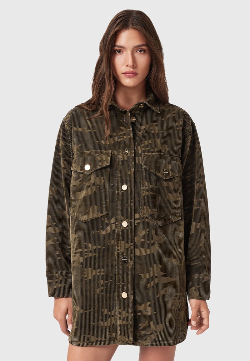 Junge Frau, die eine übergroße olivgrüne Kordjacke im Camouflage-Muster mit vorderen Knöpfen und zwei aufgesetzten Brusttaschen trägt, steht vor einem schlichten Hintergrund.