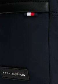 Mochila de tela azul marino con acentos de cuero negro. Presenta un parche de logo plateado y un pequeño detalle de bandera tricolor cosido.
