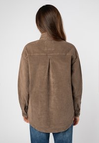 Bruine ribfluweel shirt met een rugplooitje, lange mouwen en knoopmanchetten. Versterkte schoudernaden en een zachte textuur.