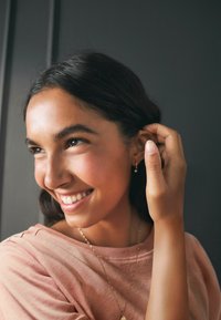 Artimos planas moters su ilgais, banguotais plaukais, dėvinčios šviesiai rožinę palaidinę ir auksinius auskarus. Švelnus apšvietimas pabrėžia jos besišypsančią veidą ir odos tekstūrą.