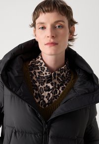 Doudoune noire avec fermeture éclair et grande capuche, superposée sur une chemise à imprimé léopard beige et un pull en tricot vert foncé.