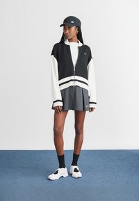 Črno-belega skrajšanega zip-up jakna z črtastim robom, siva plisejska krila, črne nogavice in bele športne čevlje. Črna kapa dopolnjuje obleko.