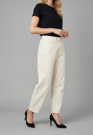 Vrouw draagt off-white rechte hoge taillebroek, zwart kort mouwen top en zwarte puntige hoge hakken, staand voor een grijze achtergrond.