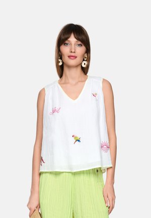 Frau trägt ein weißes ärmelloses Top mit V-Ausschnitt und bunter Stickerei sowie hellgrüne plissierte Hosen und steht vor einem einfarbigen Hintergrund.