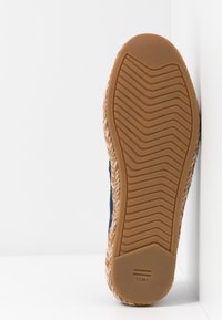 Semelle d’une chaussure marron à semelle en caoutchouc avec un motif en zigzag, marquée "TOMS,", reposant contre un coin blanc.