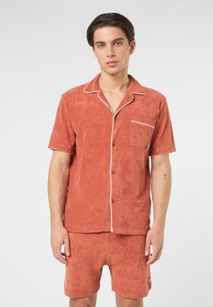 Ung mand iført rustfarvet frotté shortærmet skjorte med knapper og matchende shorts med hvide kantbånd og små logoer.